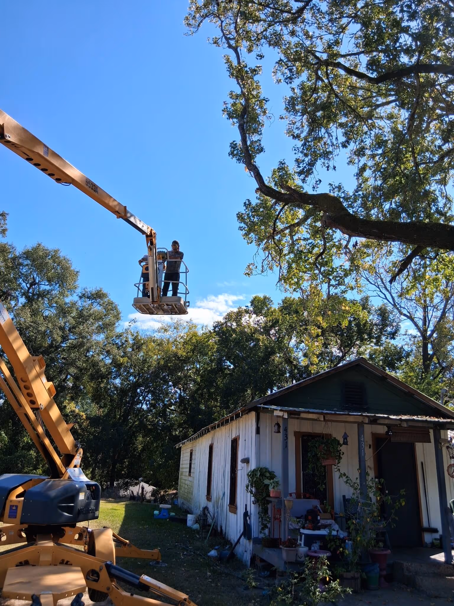 Two workers elevated on a yellow crane lift near a small white house with trees and a clear blue sky.