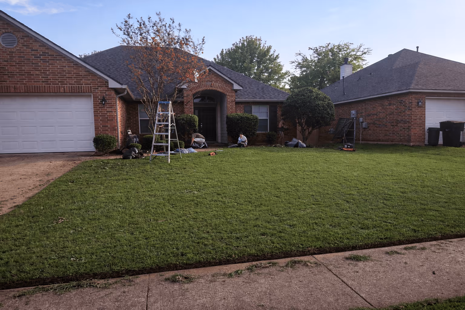Construction site with workers assembling a gray concrete block retaining wall beside a house, surrounded by trees and gravel.